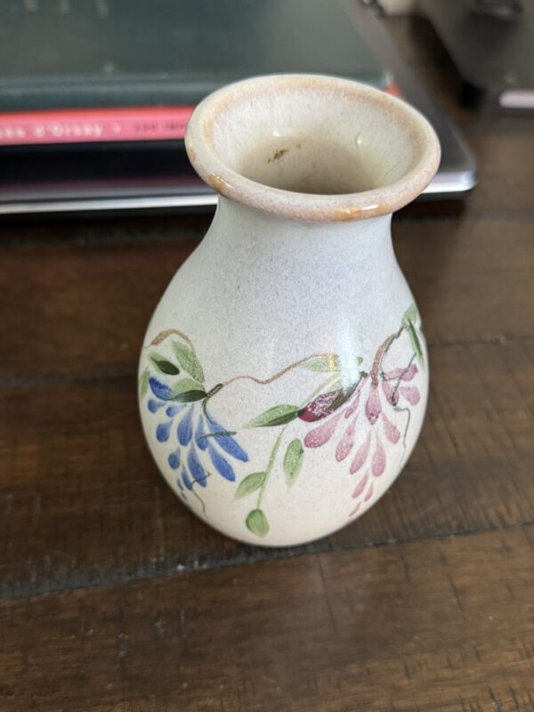 A small, round ceramic vase with hand-painted purple, blue, and green floral designs sits on a wooden surface beside a Woulind Chile Serving Dish. In the background are a closed laptop and a book.