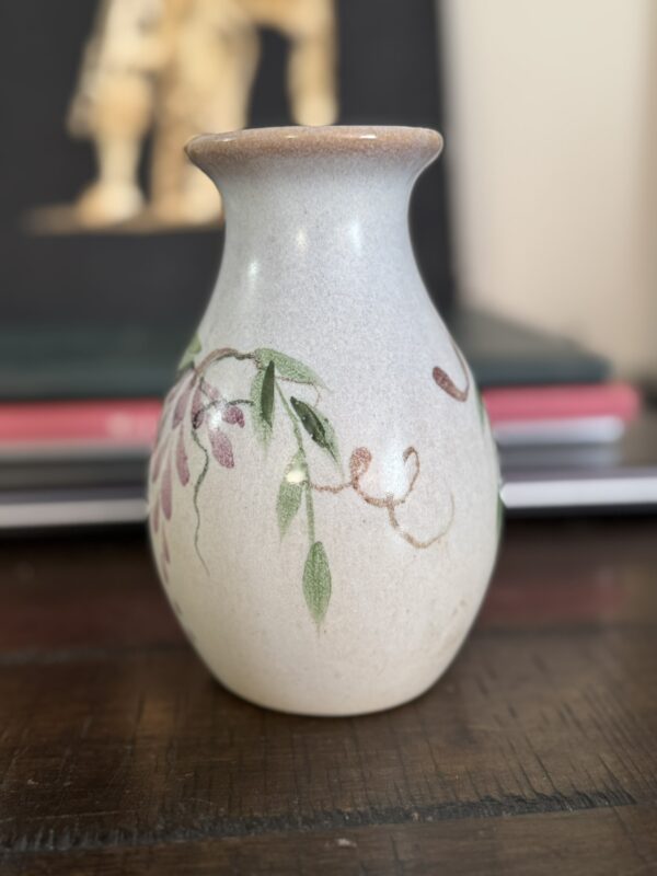 A small ceramic vase with a narrow neck, hand-painted with green leaves and pink flowers inspired by the Woulind Chile Serving Dish, sits on a dark wooden surface with blurred books and the Woulind Chile Serving Dish in the background.