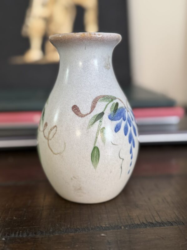 A small white ceramic vase with a brown-tinted rim and hand-painted blue flowers sits on a wooden surface beside the Woulind Chile Serving Dish. Green leaves and brown swirls decorate the vase, with blurred books in the background.