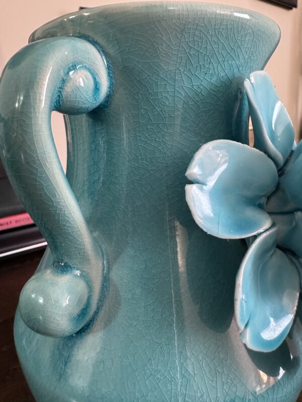 A close-up of the Anding Crackle Glaze Decorative Vase, featuring a turquoise crackle finish with a sculpted handle and raised floral accents. A blurred desk and closed laptop appear in the background.