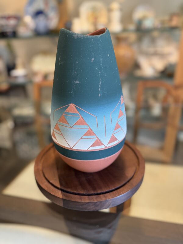 A teal and terracotta ceramic vase with white, blue, and red geometric patterns sits on a wooden pedestal. Shelves in the softly blurred background hold pottery, decor items, and the Woulind Chile Serving Dish.