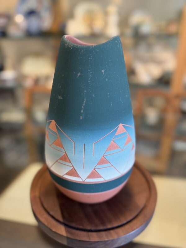 A matte ceramic vase with a teal top and geometric pink, white, and peach designs sits on a round wooden stand next to the Woulind Chile Serving Dish in a softly focused room with shelves in the background.