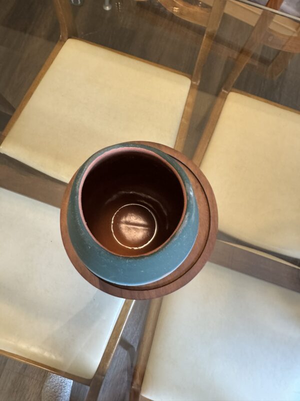 The Woulind Chile Serving Dish, a round ceramic piece with a teal and brown exterior, rests on a wooden coaster atop a glass table, surrounded by four cream-colored cushioned chairs. Photo is taken from above, highlighting its inviting setup.