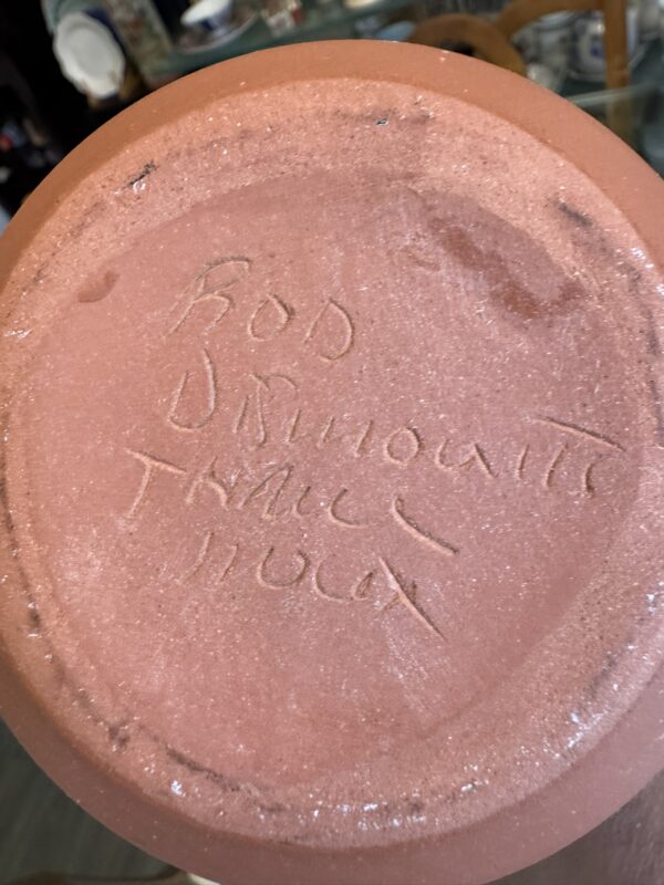 A Woulind Chile Serving Dish with “Bob Brikoutte Thack Holck” handwritten on its terracotta base. Its dusty, textured surface has white speckles and marks, while a blurred tabletop and chair appear in the background.