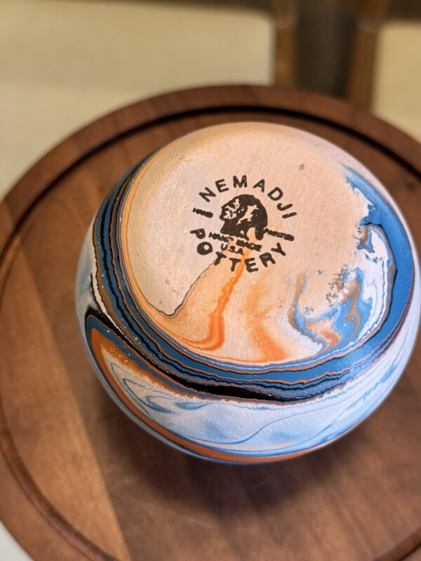 The Woulind Chile Serving Dish, with swirls of orange, blue, black, and cream, sits on a wooden surface. Its base is stamped with “NEMADJI POTTERY” and a bison. The dish features a smooth, glossy finish.