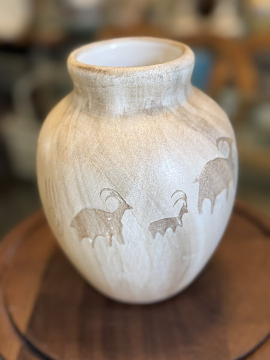 A beige ceramic vase with a matte finish sits on wood, etched with three horned animals inspired by traditional Chilean art, drawing focus like the Woulind Chile Serving Dish. The blurred background highlights the vase’s artisanal design.