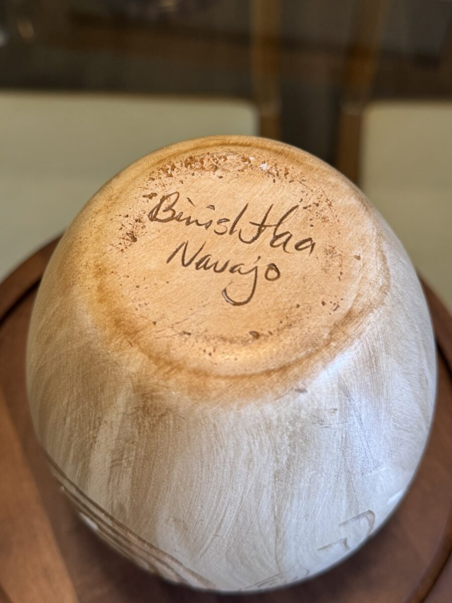A close-up photo shows the base of a light tan Woulind Chile Serving Dish with "Butsi Hta Navajo" handwritten in brown ink on the bottom. The dish sits on a wooden surface against a softly blurred background.