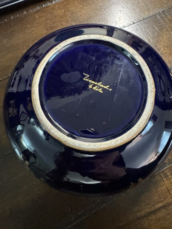 A dark blue ceramic bowl is shown upside down on a wooden surface. The bowl has a shiny glaze and a visible unglazed rim at the base. In the center, gold handwritten text reads Versailles by Gale. Reflections are visible on the glossy surface.