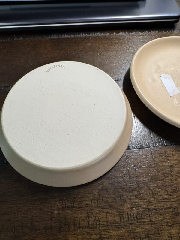 Two round, beige Woulind Chile Serving Dishes sit on a dark wooden surface—one is upside down, revealing a FELLESEN maker’s mark; the other is upright with a white rectangular sticker partially visible inside.
