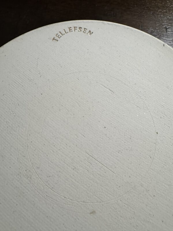 Close-up of the bottom of a round, off-white ceramic object. The surface is slightly textured with faint circular indents. The word “TELLEFSEN” is stamped in uppercase letters near the edge. The background is a dark wooden surface.
