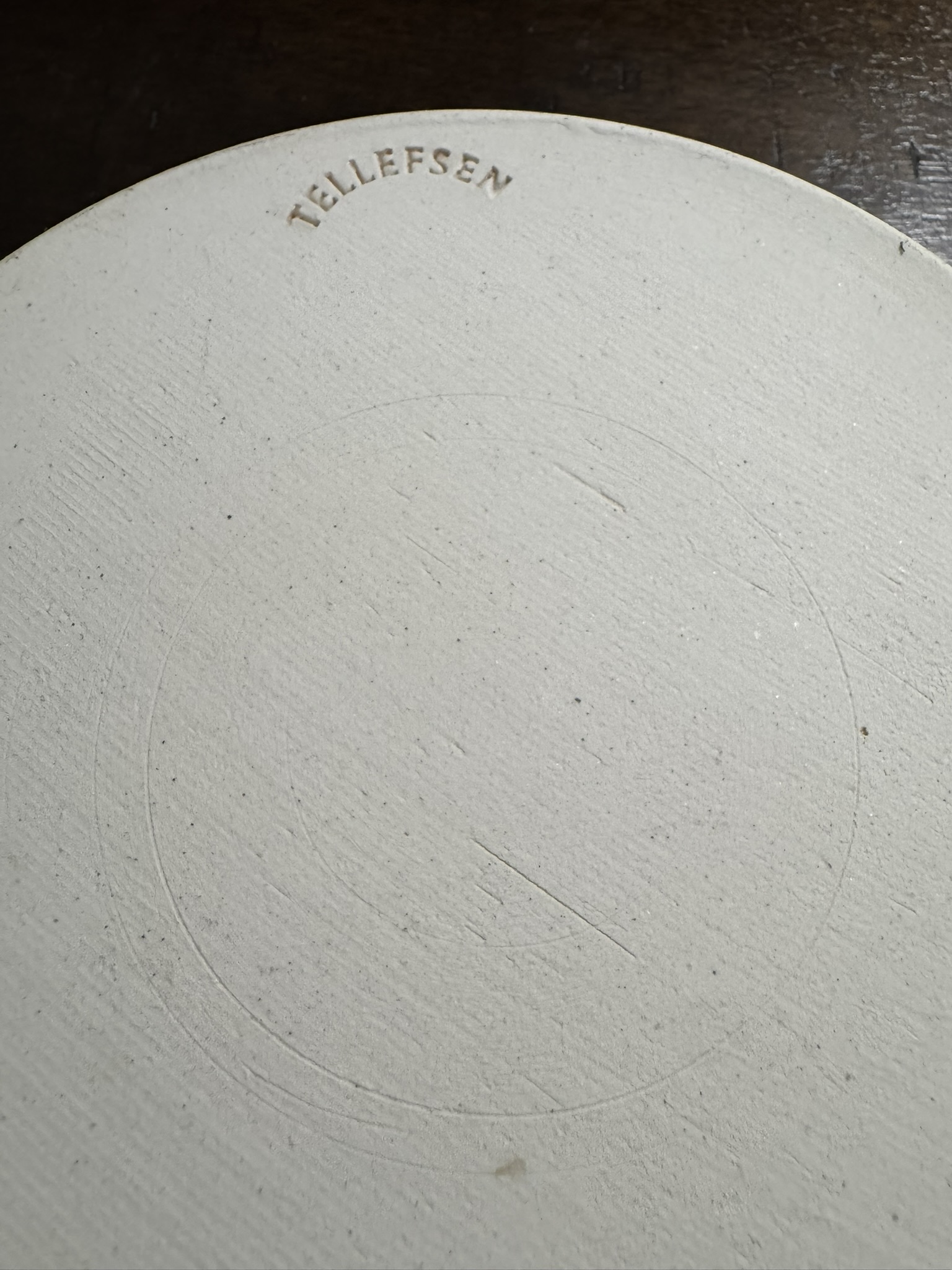 Close-up of the bottom of a round, off-white ceramic object. The surface is slightly textured with faint circular indents. The word “TELLEFSEN” is stamped in uppercase letters near the edge. The background is a dark wooden surface.