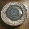 A round ceramic plate with a glazed, spiral-patterned center in shades of gray and brown. The rim is decorated with repeating scallop or fan-like embossed designs. It sits on a wooden surface.