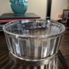 The Woulind Chile Serving Dish, featuring a clear ribbed design, sits on a wooden surface. Behind it are stacked books, a turquoise flower-patterned vase, and blurred objects like a framed photo and black lamp against a beige wall.