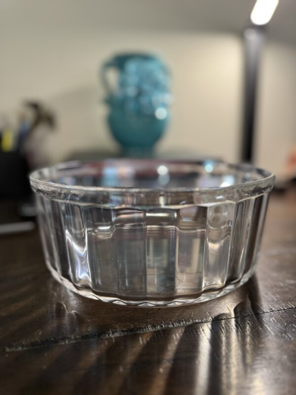 The Woulind Chile Serving Dish, made of clear glass and filled with water, sits on a dark wooden table. Light shimmers on its textured sides. In the blurred background are a turquoise vase and a black lamp, creating depth.
