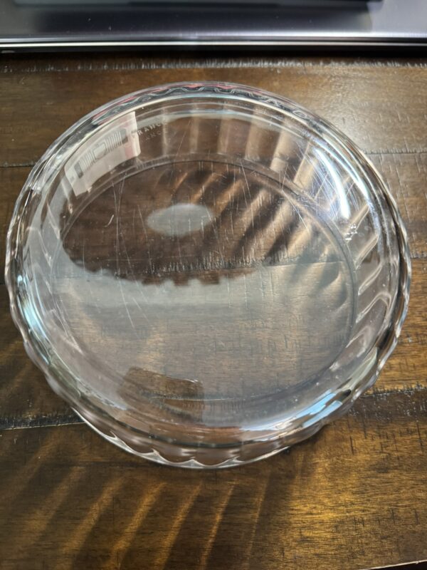 The Woulind Chile Serving Dish, made of clear glass with fluted edges, rests empty on a dark wooden table. Light reflects off its surface while a laptop appears slightly out of focus at the top of the image.