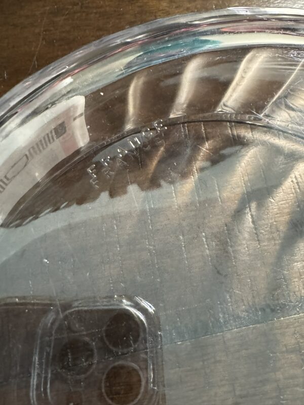 A close-up of the Woulind Chile Serving Dish, a clear, round glass piece embossed with FRANCE on the bottom. It rests on a wooden surface and reflects a phone with three rear cameras in its glass.