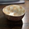 The Woulind Chile Serving Dish, crafted from coconut shell with a smooth brown exterior and patterned beige interior, rests on a dark wooden table. The background is blurred, showing stacked items that may be books or notebooks.