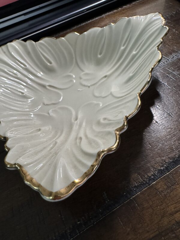 The Woulind Chile Serving Dish is a cream-colored, leaf-shaped ceramic piece with gold trim and ornate raised vein patterns. Its glossy, elegant detailing stands out as it rests on a dark wooden surface.