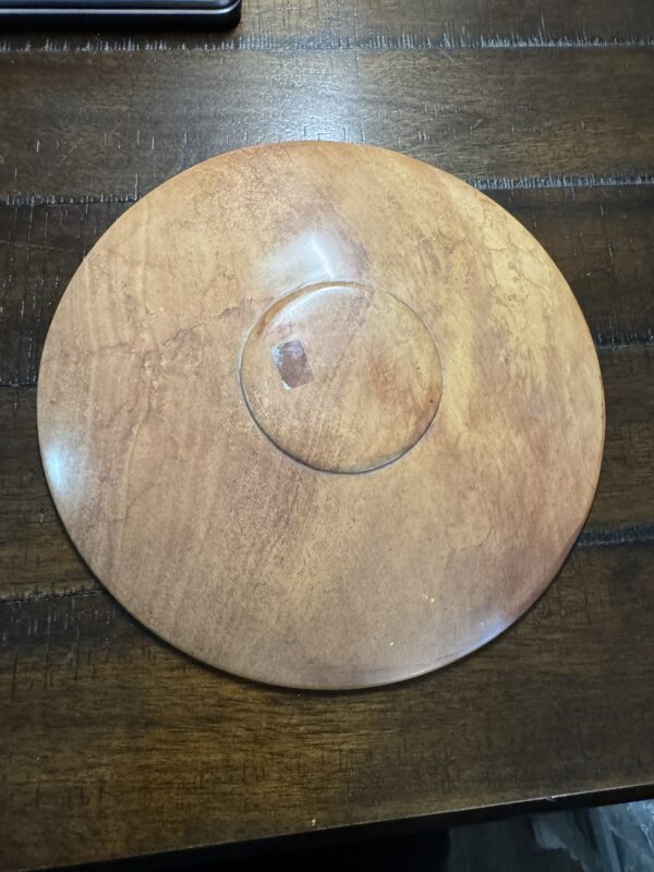 The Woulind Chile Serving Dish, round and flat with a glossy finish, rests on a dark wooden table. It features a central circular ridge and subtle wood grain patterns, resembling a wooden plate or spinning top.
