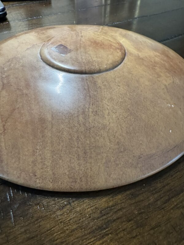 The Woulind Chile Serving Dish, a round, flat, reddish-brown piece with a raised center, sits upside-down on a dark wooden table. Its natural texture, color variations, and faint cracks suggest it's handcrafted.