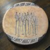 The Woulind Chile Serving Dish is a round wooden plate from Chile, engraved with eight tall, abstract tribal human figures holding staffs. Its brown, geometric-edged design makes it ideal for serving on any dark wooden surface.