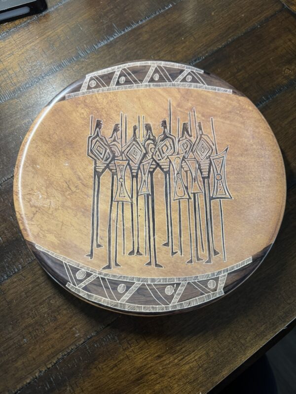 The Woulind Chile Serving Dish is a round wooden plate from Chile, engraved with eight tall, abstract tribal human figures holding staffs. Its brown, geometric-edged design makes it ideal for serving on any dark wooden surface.