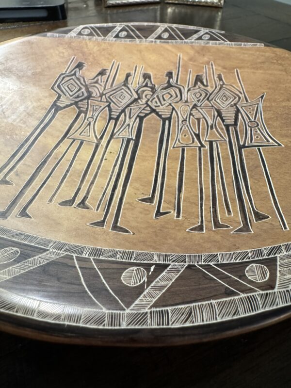 The Woulind Chile Serving Dish is a round wooden piece with carved and etched tall, thin figures holding spears in geometric patterns, and a border of triangles, circles, and lines etched along the edge.