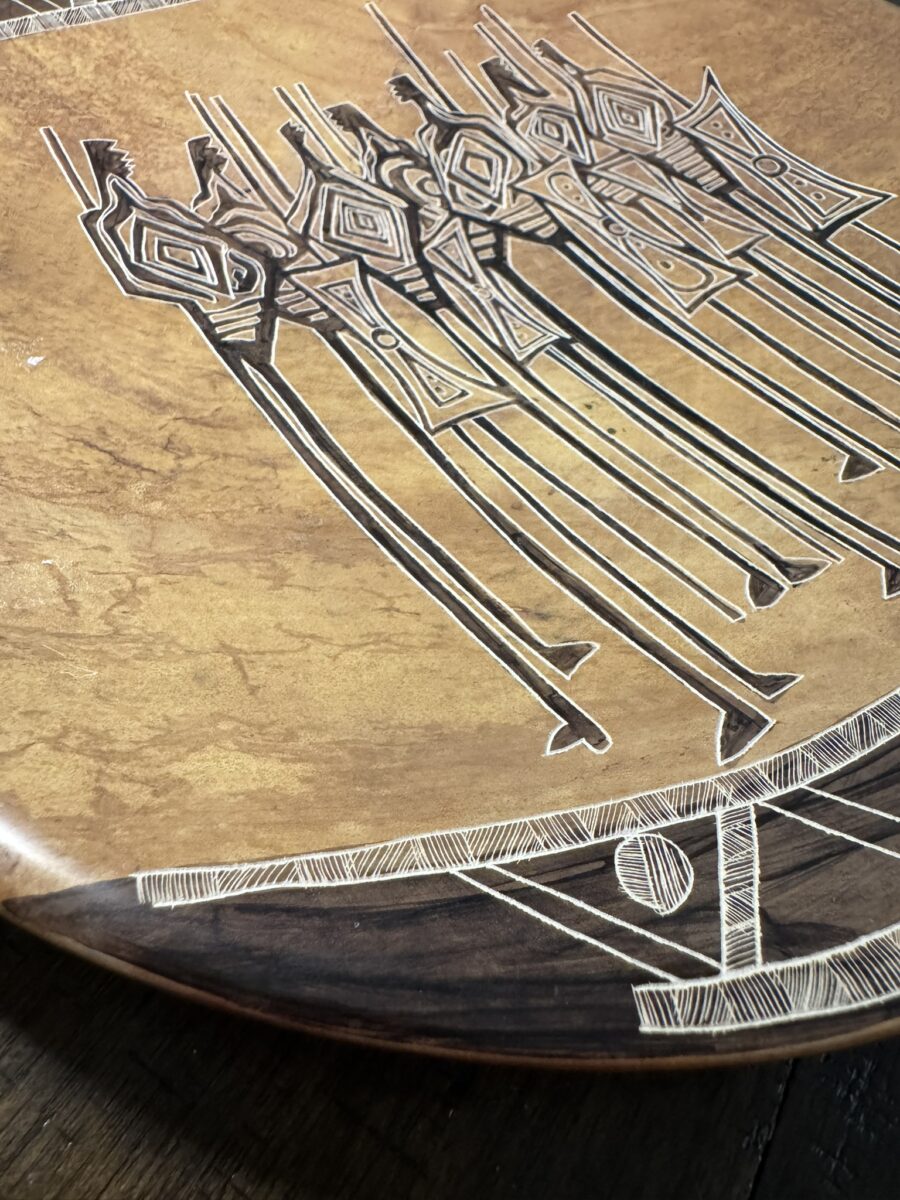 A close-up of the Woulind Chile Serving Dish—an elegant wooden plate featuring stylized human figures with spears and bold geometric patterns etched along its detailed borders, showcasing the vibrant wood grain.