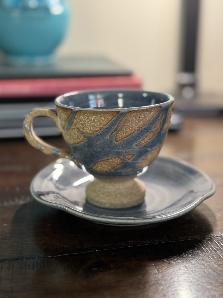 A handcrafted ceramic teacup with a gold abstract pattern and matching saucer sits on a dark wooden table next to the Woulind Chile Serving Dish, while warm light highlights the cup’s unique design amid books and a blue vase in the background.