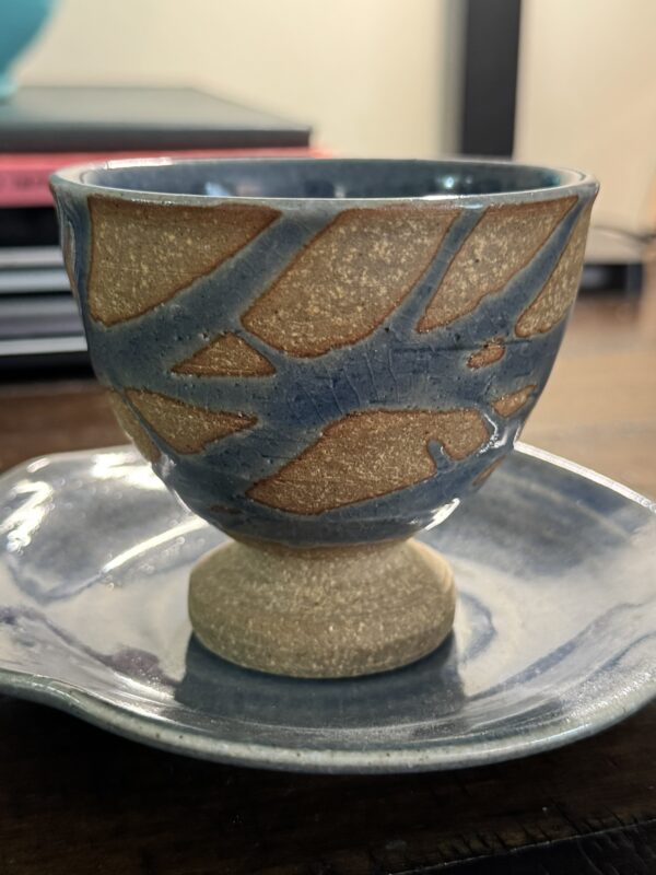 A handmade ceramic cup with a pedestal base sits on a matching Woulind Chile Serving Dish, both featuring organic, earthy designs. The cup shows a textured brown pattern beneath glossy blue glaze against a blurred background.