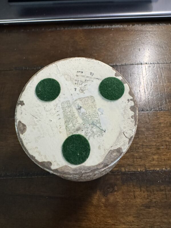 The Woulind Chile Serving Dish features a round, weathered design with a faded white bottom, resting on three green felt pads arranged in a triangle. The surface displays worn spots and remnants of old stickers or markings.
