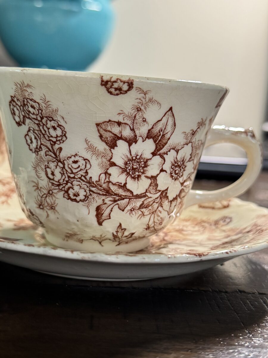 A vintage teacup and saucer with brown floral patterns on cream porcelain sits on a dark wooden surface. In the background, a blurred Woulind Chile Serving Dish in turquoise adds a pop of color. The teacup’s handle and details are in sharp focus.