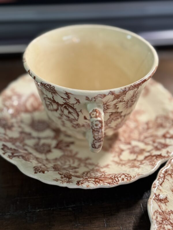 The Woulind Chile Serving Dish, a cream teacup and saucer with brown floral patterns, sits on a dark wooden surface. Its ornate handle and rim stand out against the softly blurred background, highlighting its classic elegance.