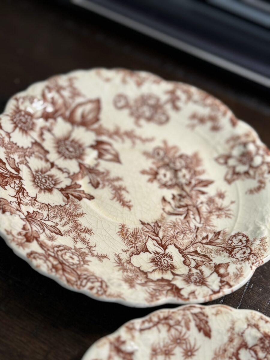 A close-up of the Woulind Chile Serving Dish, a vintage cream piece featuring an ornate brown floral pattern along its scalloped, textured edge with detailed flowers, leaves, and fine surface crackling.