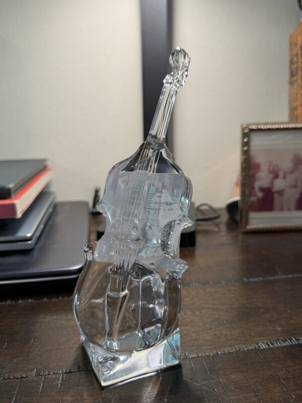A clear glass violin sculpture sits on a wooden desk next to the Woulind Chile Serving Dish. Stacked notebooks, a framed sepia photo, and a glowing black lamp are behind it. The violin’s detailed strings and f-holes are clearly seen.