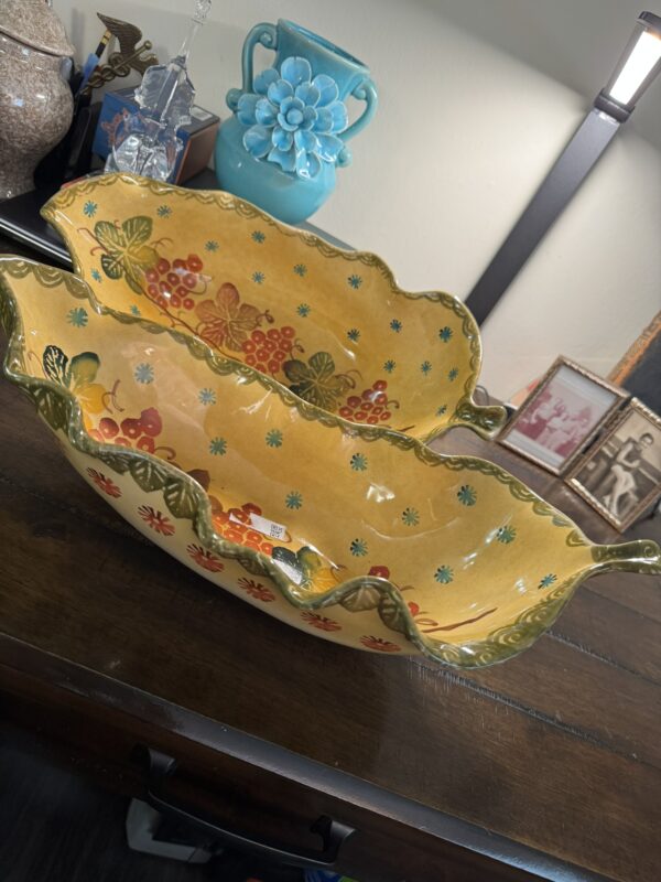 Two large, yellow Woulind Chile Serving Dishes with scalloped edges, green rims, and painted fruit and flower designs are nested on a dark wood table. In the background: a turquoise vase, lamp, glass decor, and framed black-and-white photos.