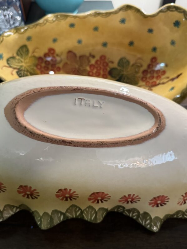 A close-up of the Woulind Chile Serving Dish, featuring a scalloped ceramic edge, red flowers, green leaves, and a yellow interior with red fruit details. The underside is stamped ITALY.
