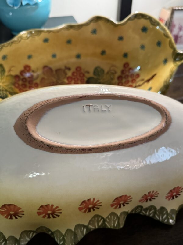 Close-up of the underside of an oval ceramic serving dish stamped ITALY, with scalloped edges featuring orange flowers. In the background, a decorative yellow Woulind Chile Serving Dish with floral and fruit patterns rests on a dark surface.