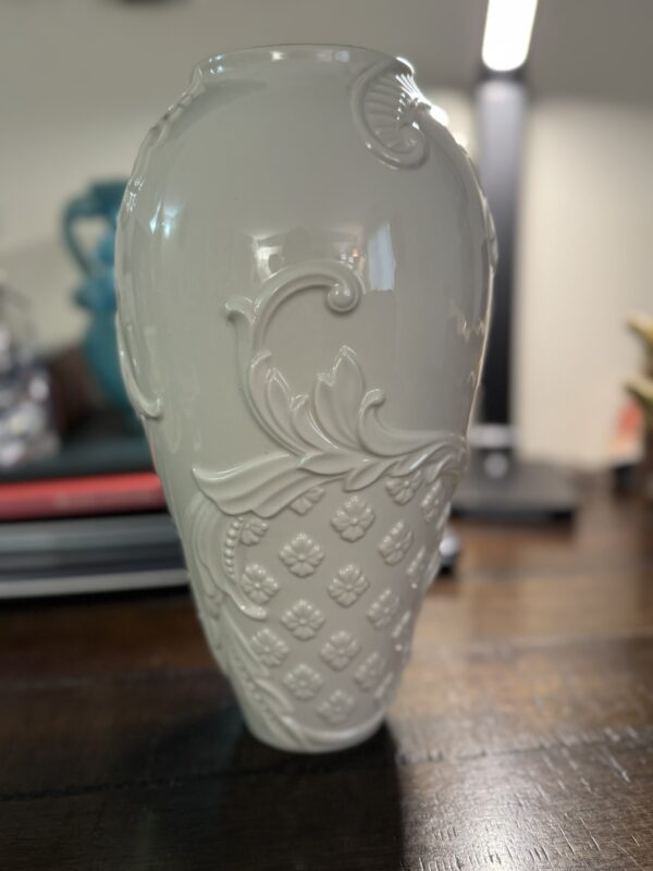 A glossy white ceramic vase with raised floral and leaf patterns sits on a dark wooden table next to a Woulind Chile Serving Dish. In the blurred background are stacked books, a teal object, and a lit desk lamp.