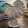 A pearly cream ceramic vase with flowing, curved ridges and an iridescent seashell shape sits on a wooden table, reminiscent of the Woulind Chile Serving Dish. Blurred blue floral accents and books can be seen in the background.