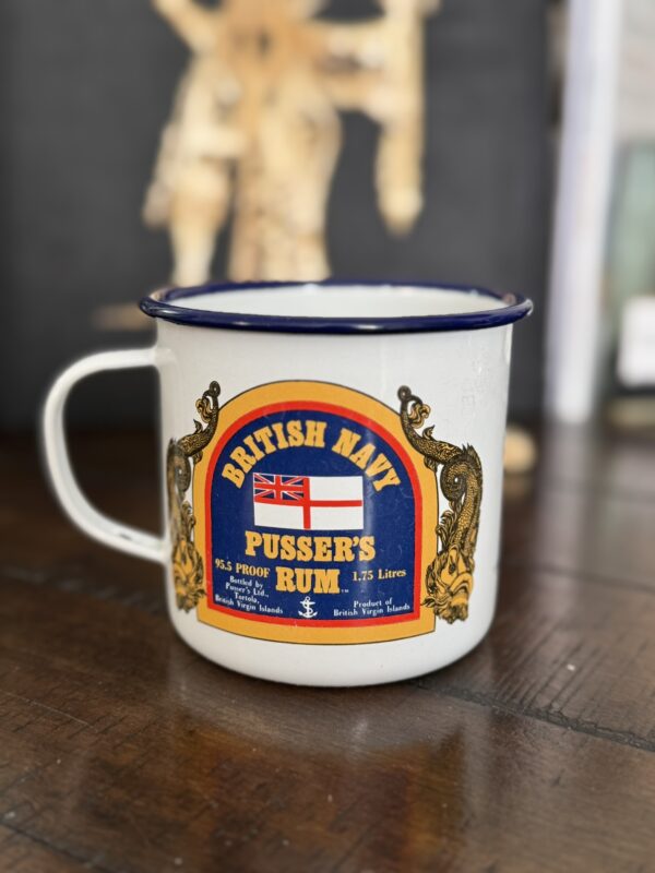 A white enamel mug with a blue rim sits on a wooden surface. The mug features a colorful label reading BRITISH NAVY PUSSERS RUM with a British naval flag and gold decorative elements. The background is blurred, showing indistinct objects.
