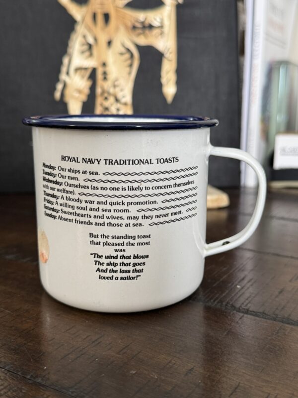 A white enamel mug with a blue rim sits on a wooden surface. Printed on the mug are ROYAL NAVY TRADITIONAL TOASTS for each day of the week, along with a short poem about a ship and a sailor. A blurred golden ornament and books are in the background.