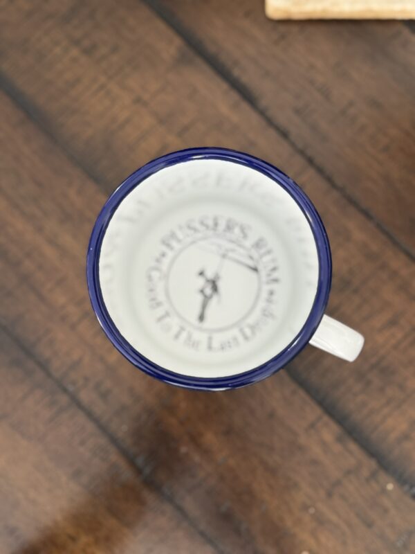 A white enamel mug with a dark blue rim sits on a wooden surface. Inside the mug, the base has a logo and text: Pusser’s Rum with a sailor holding a rope, and Gods in the Last Drop in smaller print. The image is taken from above.