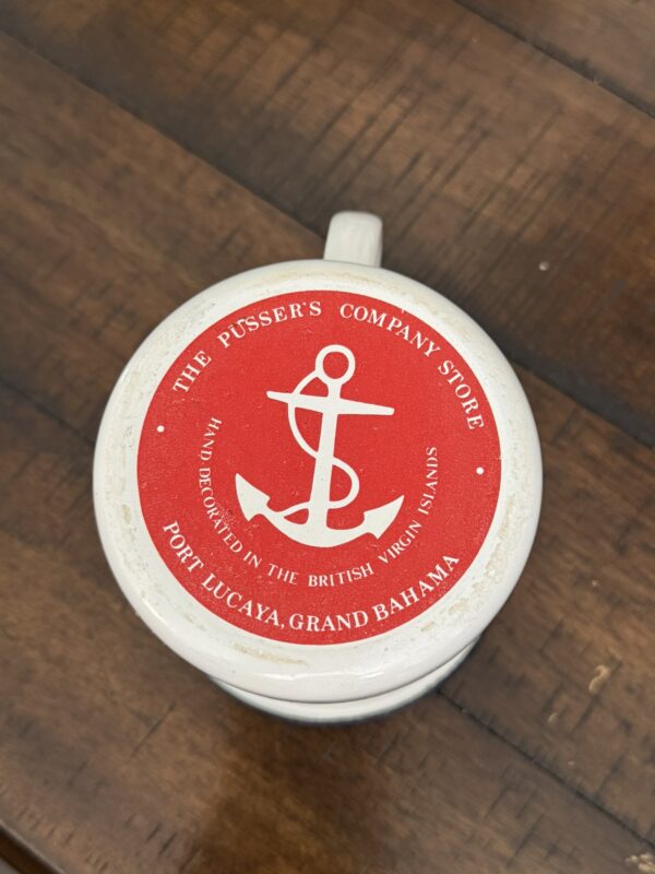 A white mug sits on a wooden surface, viewed from above. The mug’s lid displays a red circle with a white anchor in the center, surrounded by text reading: “THE PUSSERS COMPANY STORE PORT LUCAYA, GRAND BAHAMA HAND DECORATED IN THE BRITISH VIRGIN ISLANDS.”.