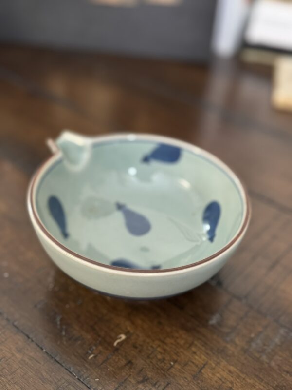A small ceramic bowl with a spout sits on a wooden table. The bowl is pale green with a brown rim and features dark blue, teardrop-shaped patterns on the inside wall. The background is blurred, focusing attention on the bowl.