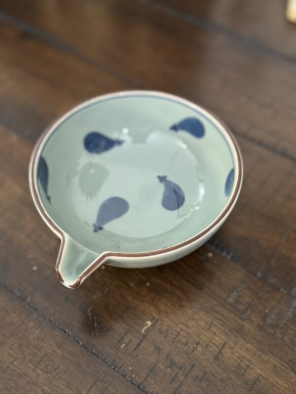 A small, round ceramic bowl with a pouring spout sits on a wooden surface. The bowl is light green with a brown rim and features blue balloon or drop-shaped patterns on its inner surface. The wood beneath shows textured grain and lines.