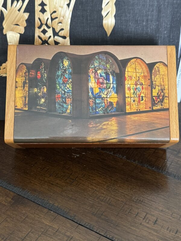 A wooden box with a lid decorated by a photo of arched stained-glass windows. The colorful windows depict various abstract designs and figures, lit from behind by sunlight. The box rests on a dark wooden surface with a black and gold decorative background.