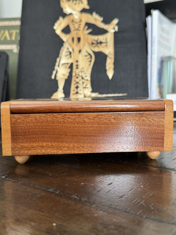 A rectangular wooden box with rounded corners and small knob feet sits on a dark wood surface. Behind it, there is an artwork depicting a gold silhouette of a traditional figure on a black background. Other books and papers are visible in the background.