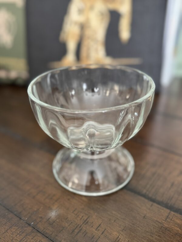 A clear, empty glass dessert bowl with a fluted design sits on a dark wooden surface. The bowl has a wide rim and a sturdy pedestal base. The background is softly blurred, showing hints of artwork and books.