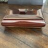 A small, rectangular wooden box with a sloped lid and a round knob sits on a wooden table. The box has a polished, dark brown finish, and blurred objects can be seen in the background.